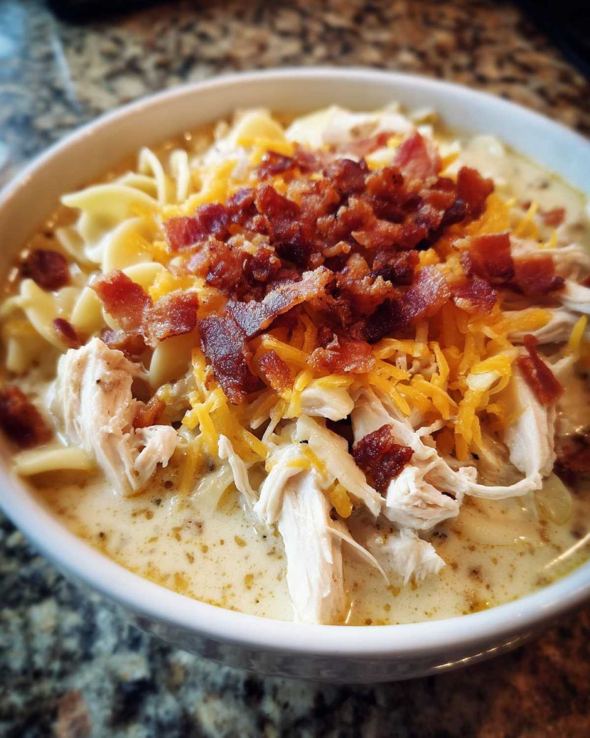 A close-up of a hearty bowl of creamy Crack Chicken Noodle Soup topped with shredded chicken, cheddar cheese, and crispy bacon bits.