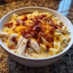 A white bowl filled with creamy Crack Chicken Noodle Soup, topped with shredded chicken, cheddar cheese, and crispy bacon bits.