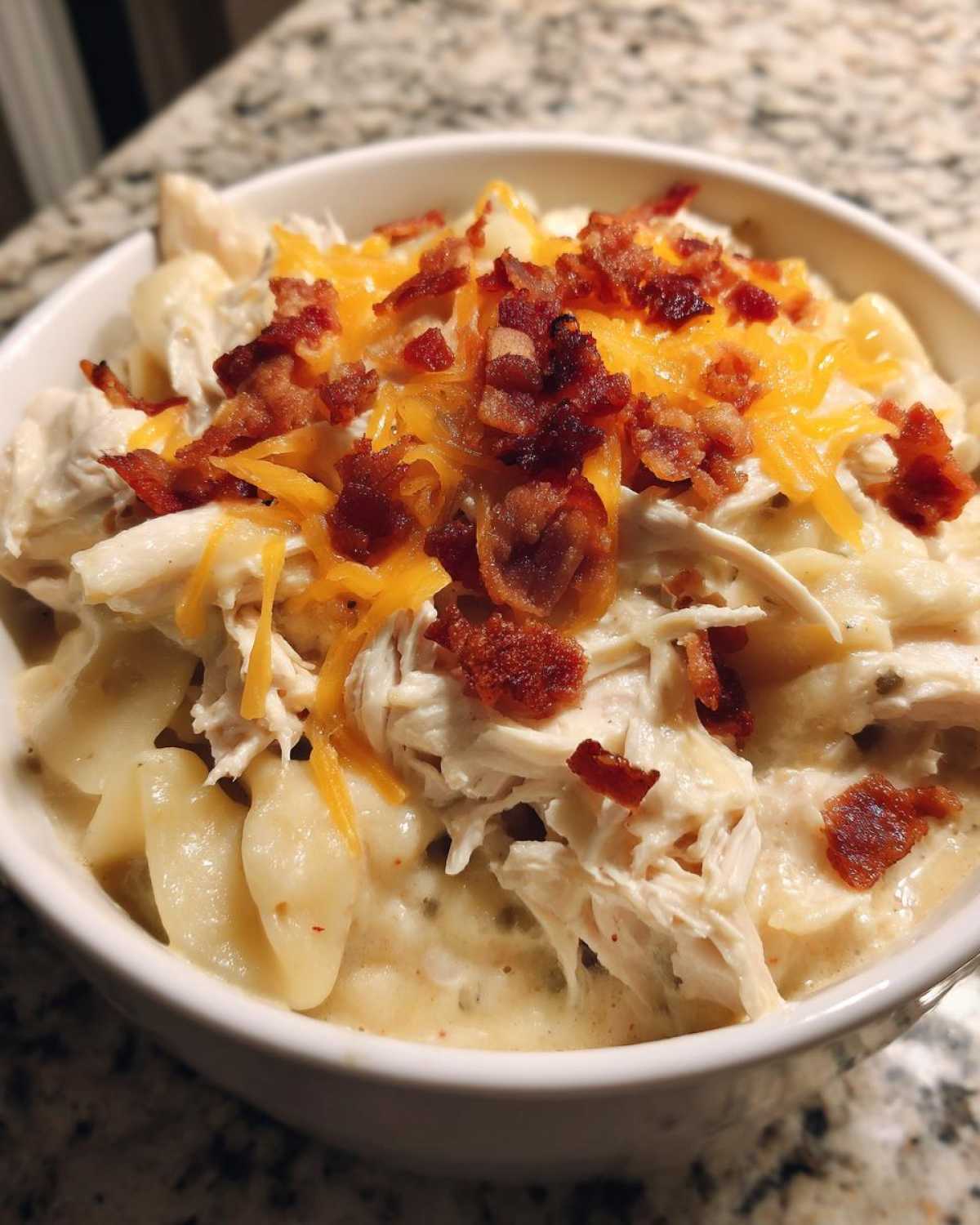 A close-up of a creamy bowl of Crack Chicken Noodle Soup topped with shredded chicken, cheddar cheese, and crispy bacon bits.