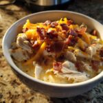 A close-up of a white bowl filled with creamy Crack Chicken Noodle Soup, topped with shredded chicken, cheddar cheese, and bacon bits.
