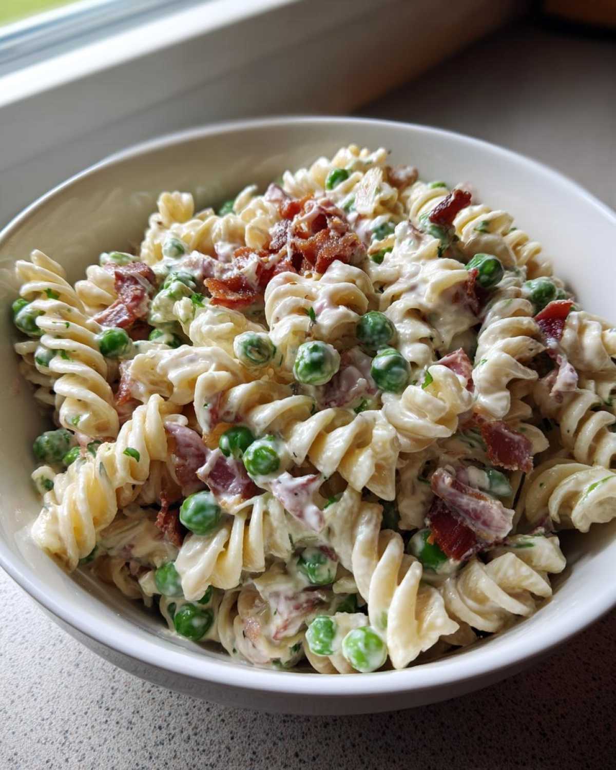 Close-up of a white bowl filled with creamy Crack Pasta Salad featuring rotini pasta, green peas, and crispy bacon pieces.
