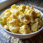 A close-up of creamy Hellman's Potato Salad mixed with chunks of egg and green onions, served in a white bowl.