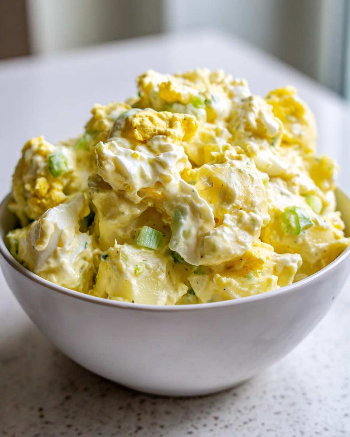 A close-up of creamy Hellman's Potato Salad mixed with chunks of egg and green onions in a white bowl.