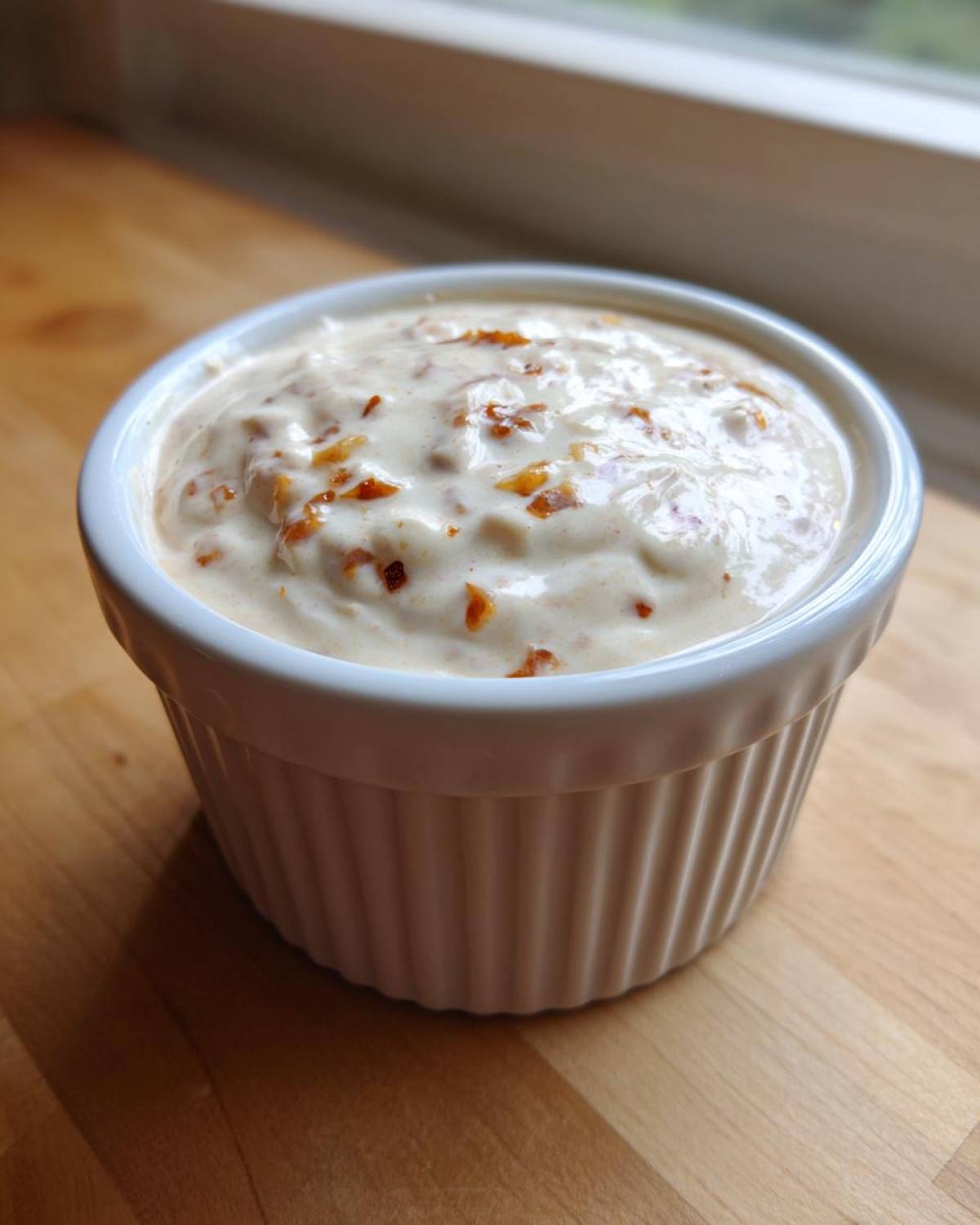 Close-up of creamy Hot Honey Ranch Sauce mixed with visible flakes of chili or honey solids in a white fluted ramekin.