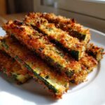 A stack of golden brown, crispy baked parmesan zucchini sticks piled high on a white plate.