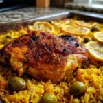 Close-up of a perfectly cooked, crispy-skinned chicken thigh atop yellow rice with olives and lemon slices in a baking pan, showcasing the Crispy One Pan Portuguese Chicken and Rice Bake.