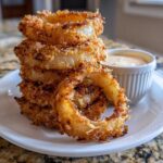 A tall stack of golden brown, crispy Tennessee Onions rings served on a white plate next to a small ramekin of dipping sauce.