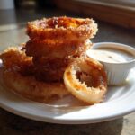A stack of golden brown, crispy Tennessee Onions Rings served on a white plate next to a small cup of dipping sauce.