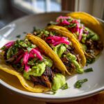 Close-up of three delicious Mushroom Tacos loaded with dark filling, bright green avocado crema, and pink pickled onions.