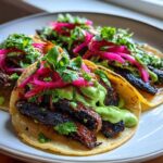 Three close-up Mushroom Tacos filled with dark, savory mushrooms, drizzled with green sauce, and topped with bright pink pickled onions and cilantro.