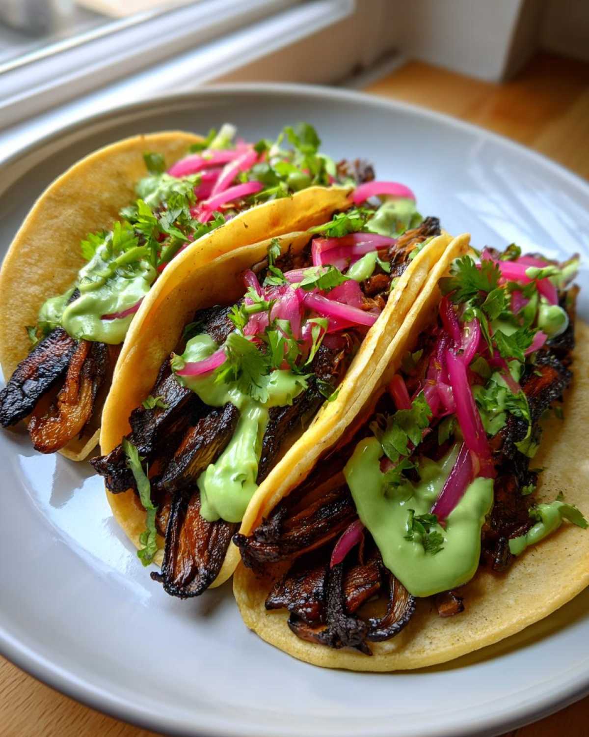 Close-up of three delicious Mushroom Tacos filled with dark, savory mushrooms, topped with bright pink pickled onions, cilantro, and a creamy green sauce.