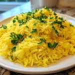 A mound of bright yellow Greek Lemon Rice topped with fresh chopped parsley on a white plate.
