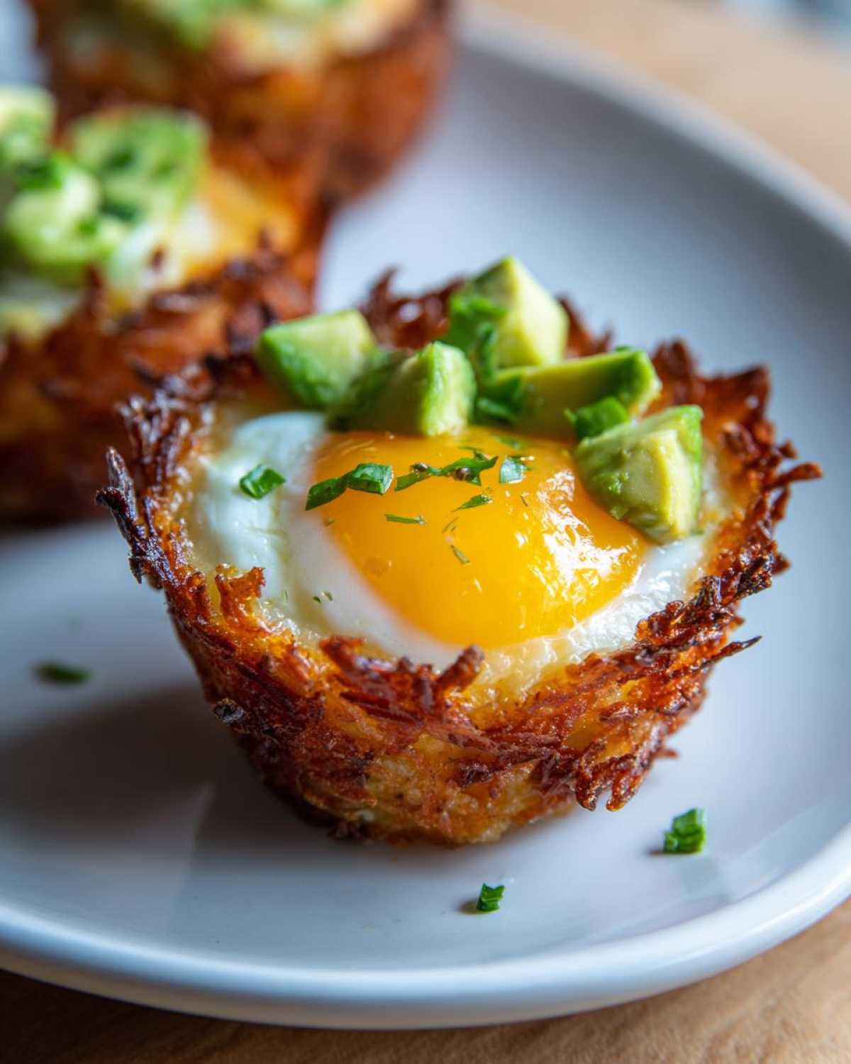 A close-up of a crispy Hash Brown Avocado Egg Cup featuring a bright yellow sunny-side-up egg topped with diced avocado.