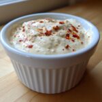 Close-up of creamy Hot Honey Ranch Sauce in a white ramekin, topped with visible red chili flakes.