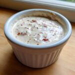A close-up view of creamy Hot Honey Ranch Sauce speckled with red pepper flakes and herbs in a small white ramekin.