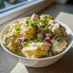 A close-up of creamy Jamaican Jerk Potato Salad mixed with red onion and topped with green onions in a white bowl.