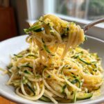 A fork lifting a generous portion of Lemon Zucchini Pasta, topped with bright lemon zest and zucchini ribbons.