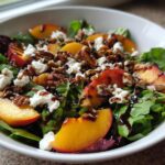 A vibrant close-up of a Peach Feta Salad featuring fresh greens, sliced peaches, crumbled feta, pecans, and a balsamic glaze.