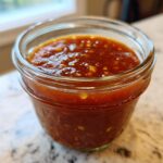 Close-up of homemade Pineapple Jalapeno Bourbon BBQ Sauce stored in a small glass jar on a granite countertop.
