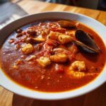 A close-up of a steaming bowl of rich, red tomato-based Cioppino filled with shrimp, white fish chunks, and mussels.