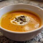 A close-up of a bowl of vibrant orange Roasted Butternut Squash Soup, garnished with cream swirl and pumpkin seeds.