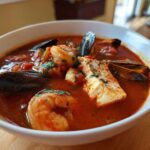 Close-up of a white bowl filled with rich, red tomato broth Cioppino, featuring shrimp, mussels, and a white fish fillet.