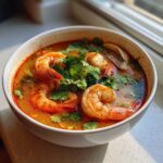 A close-up, appetizing photo of a bowl of rich, orange-red Thai Shrimp Soup topped with large cooked shrimp and fresh cilantro.