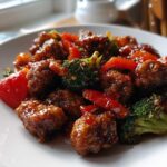 A close-up of glazed, saucy pieces of Thai Chicken Sweet and Spicy mixed with bright red bell peppers and green broccoli florets on a white plate.