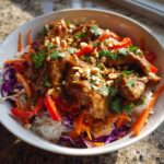 A close-up of a vibrant Thai Peanut Chicken Bowl featuring saucy chicken over rice, topped with shredded carrots, red cabbage, and crushed peanuts.