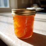 A glass jar filled with bright orange Apricot Jam with Vanilla and Lemon, sitting on a sunlit countertop.
