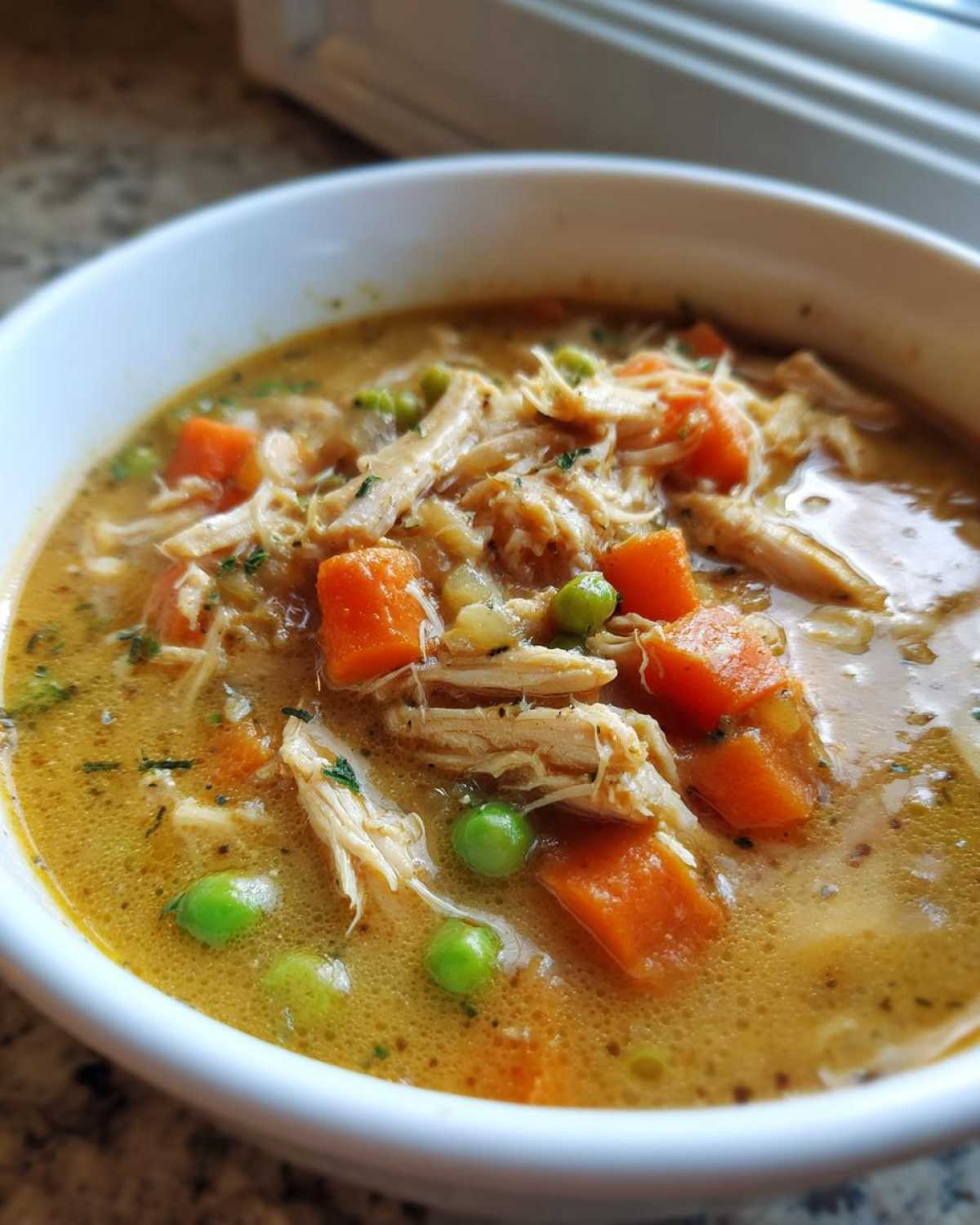 Close-up of a bowl of creamy Chicken Pot Pie Soup, featuring shredded chicken, bright orange carrots, and green peas.