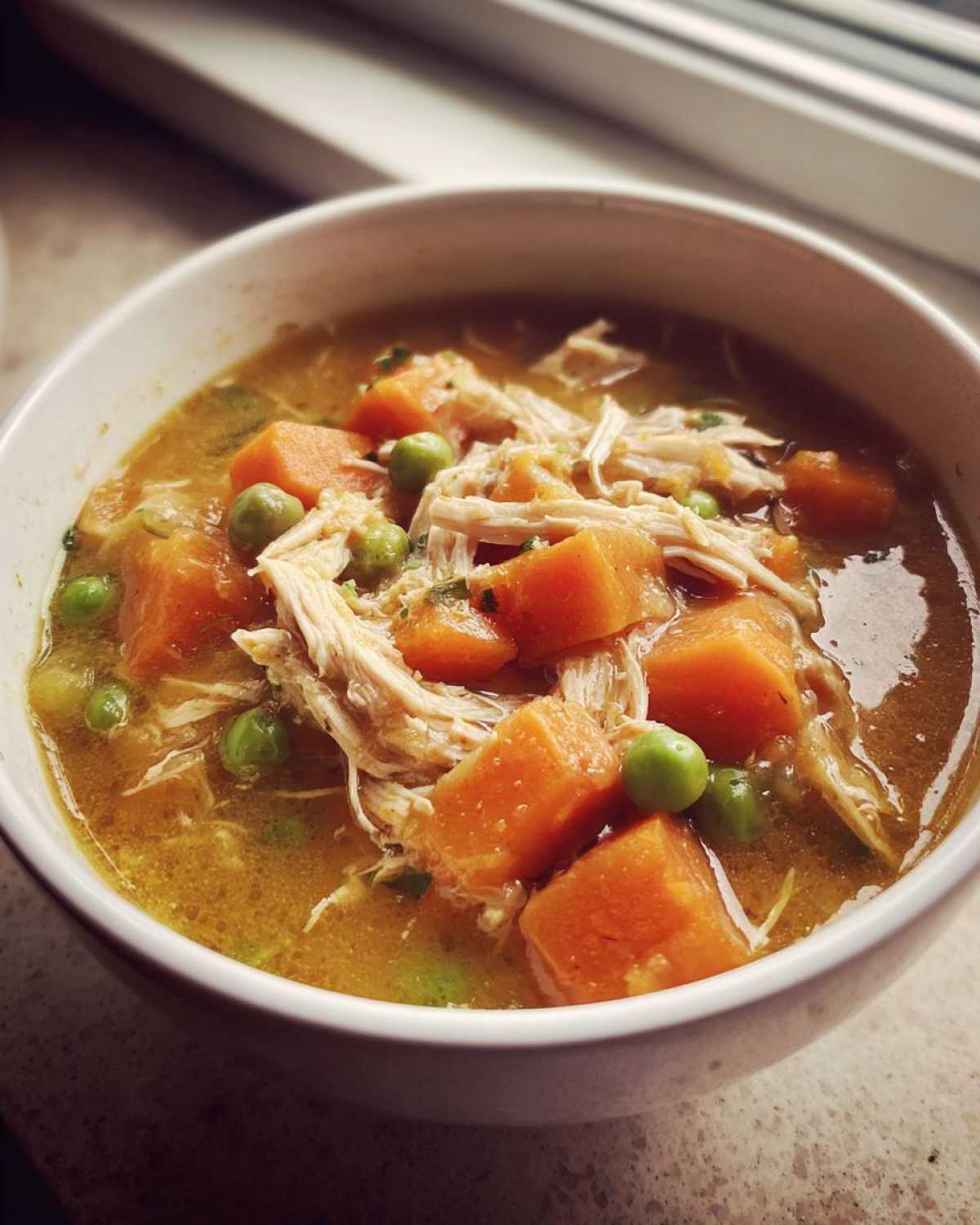 Close-up of a white bowl filled with savory Chicken Pot Pie Soup, featuring shredded chicken, large chunks of orange carrots, and green peas.