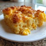 A square serving of creamy Corn & Squash Casserole with a golden, slightly browned cheesy top, served on a white plate.