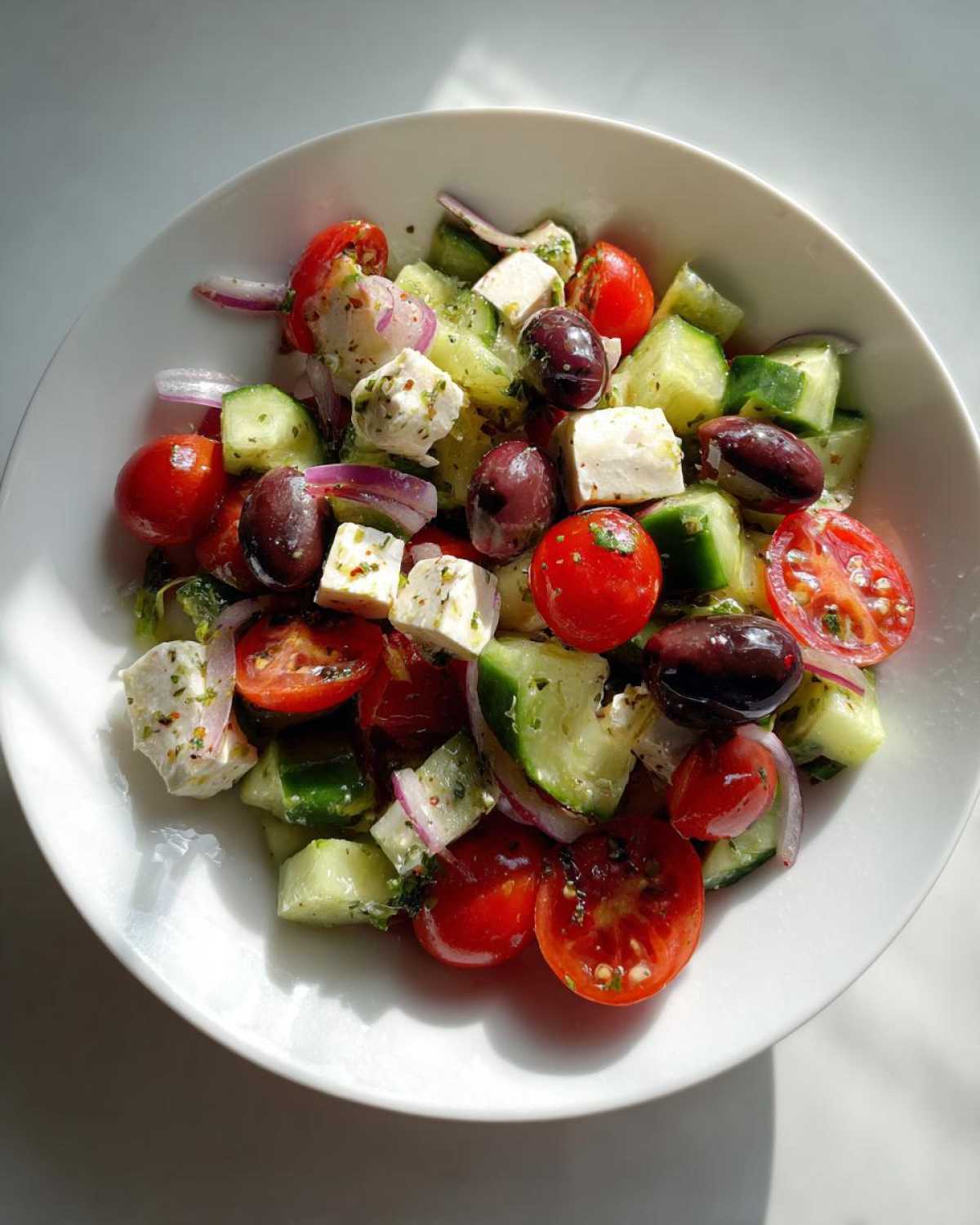 Overhead view of a bowl filled with ingredients resembling a Greek Pasta Salad: cherry tomatoes, cucumbers, feta, and olives.