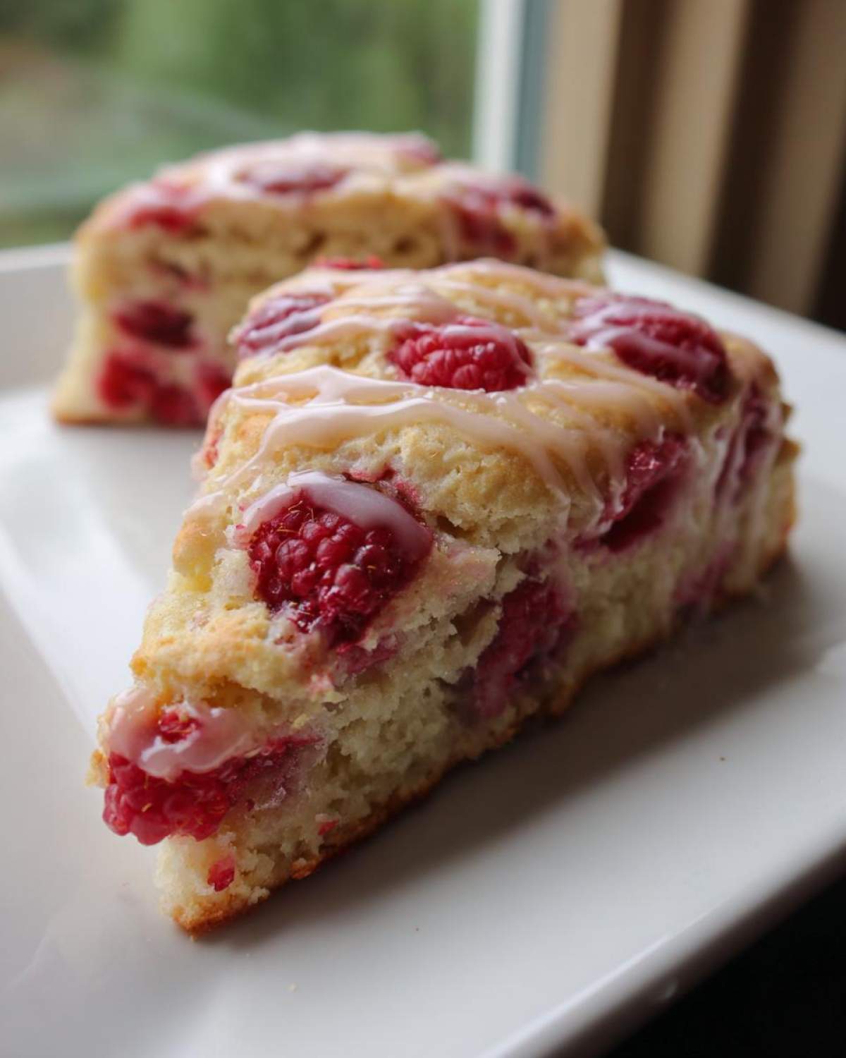 A close-up of a freshly baked Lemon Raspberry Scone, topped with a light glaze and studded with bright red raspberries.