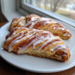 Two golden, flaky Rhubarb Danish Twists drizzled with white icing, resting on a white plate near a window.