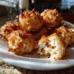 A pile of golden-brown, toasted Crab Bombs on a white plate, one is broken open showing the shredded interior.