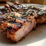 Close-up of a juicy, thick Grilled Rosemary Pork Chop showing a slight pink center and charred rosemary seasoning.