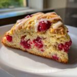 A single slice of a fluffy Lemon Raspberry Scones, topped with bright red raspberries and a white glaze, sitting on a white plate.