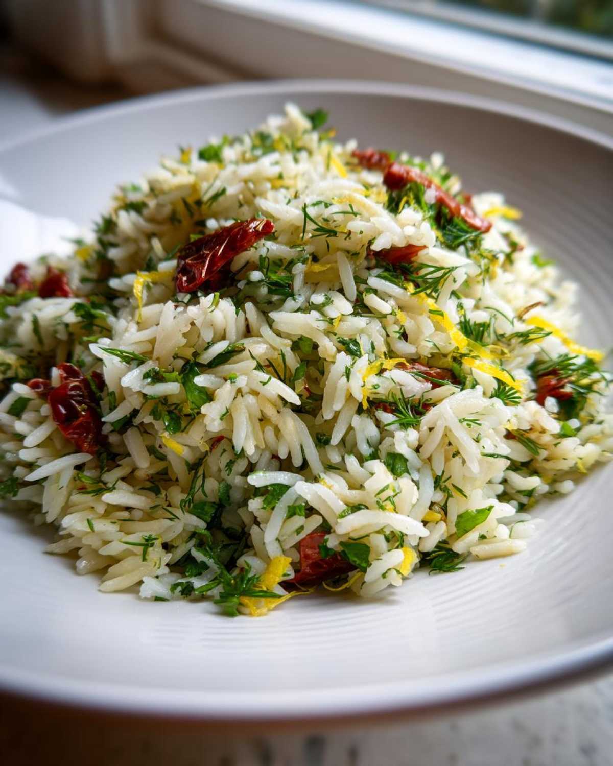 Close-up of fluffy Mediterranean Rice mixed with fresh dill, parsley, lemon zest, and sun-dried tomatoes in a white bowl.