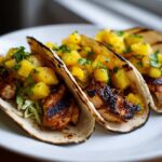 Three grilled Pineapple Chicken Tacos served on soft tortillas with pineapple salsa and shredded cabbage on a white plate.