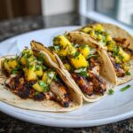 Three grilled Pineapple Chicken Tacos loaded with charred chicken and fresh pineapple salsa on a white plate.