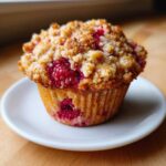 A close-up of a freshly baked Raspberry Streusel Muffin topped with golden brown crumb topping, sitting on a small white plate.